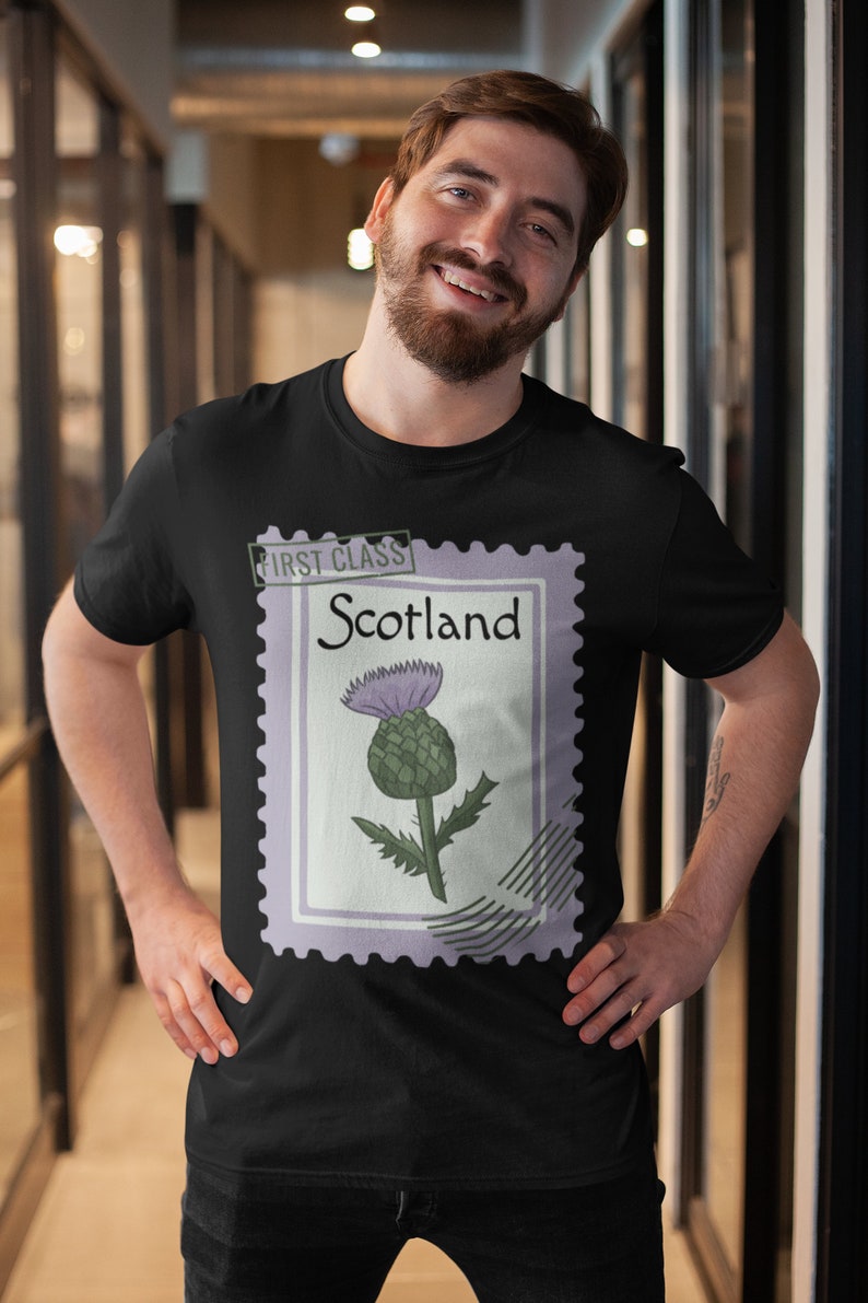 May include: Black t-shirt with a postage stamp design. The stamp shows a thistle and the words "First Class" and "Scotland". The thistle is purple and green. The t-shirt is worn by a person.
