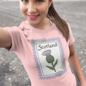 May include: Pale pink t-shirt with a postage stamp design. The stamp features the word "Scotland" and a thistle illustration. The stamp is bordered in lavender and has the words "First Class" at the top.