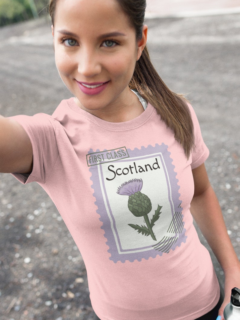 May include: Pale pink t-shirt with a postage stamp design. The stamp features the word "Scotland" and a thistle illustration. The stamp is bordered in lavender and has the words "First Class" at the top.