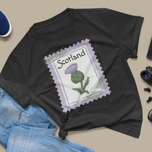 May include: Black t-shirt with a Scotland stamp design featuring a thistle illustration. The stamp is bordered in lavender and has the words "First Class" and "Scotland" printed on it. The shirt is styled with blue jeans and trainers.
