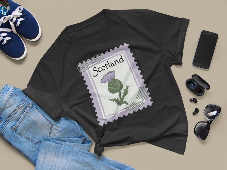 May include: Black t-shirt with a Scotland stamp design featuring a thistle illustration. The stamp is bordered in lavender and has the words "First Class" and "Scotland" printed on it. The shirt is styled with blue jeans and trainers.