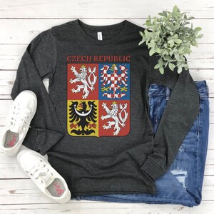 May include: Dark gray long-sleeve shirt with the text "Czech Republic" above a colorful emblem. The emblem features a coat of arms with a white lion, a checkered eagle, and other heraldic symbols. The shirt is paired with blue jeans and white sneakers.