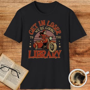 May include: Black t-shirt with a vintage-style graphic. The design features a woman riding a motorcycle with a sidecar, and the text "GET IN LOSER WE'RE GOING TO THE LIBRARY" in red and brown.