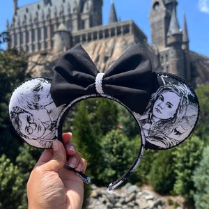 Puede incluir: Orejas de Minnie Mouse de Harry Potter en blanco y negro con un lazo negro. Las orejas presentan imágenes de Harry Potter y Hermione Granger. Las orejas están delante de un edificio que se parece a Hogwarts.