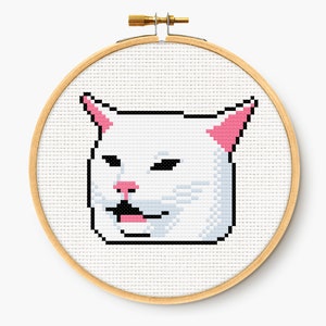 May include: A cross-stitch embroidery hoop featuring a pixelated image of a white cat with pink ears and nose, and a black mouth. The cat is set against a white background, and the hoop is made of light-colored wood.
