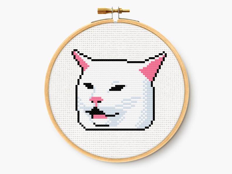 May include: A cross-stitch embroidery hoop featuring a pixelated image of a white cat with pink ears and nose, and a black mouth. The cat is set against a white background, and the hoop is made of light-colored wood.