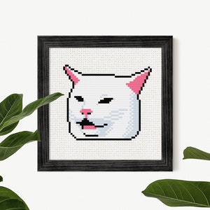 May include: Cross-stitch art of the Smudge the Cat meme, framed in a black wooden frame. The cat is white with pink accents on the ears and nose, and a black outline. The cat's mouth is open, with a pink tongue visible.