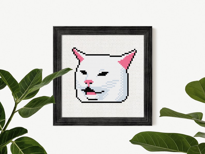 May include: Cross-stitch art of the Smudge the Cat meme, framed in a black wooden frame. The cat is white with pink accents on the ears and nose, and a black outline. The cat's mouth is open, with a pink tongue visible.
