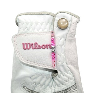 May include: White golf glove with a pink Wilson logo and a pink and silver beaded strap. The glove has a white button with a pearl-like center.