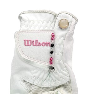 May include: White golf glove with the pink "Wilson" logo. The glove has a button closure and decorative pink and black beads. The glove is made of a textured material and has a mesh panel.