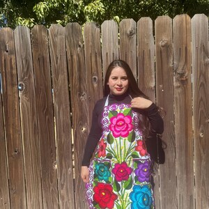 May include: A white apron with colorful embroidered flowers and a purple trim. The apron has two pockets and a white strap around the neck. The model is wearing a black long-sleeved shirt. "Model 7" is written in black text at the bottom of the image.