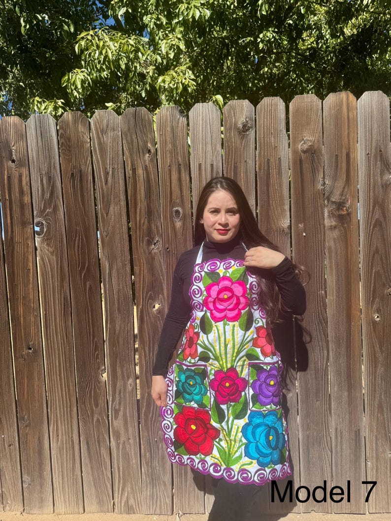 May include: A white apron with colorful embroidered flowers and a purple trim. The apron has two pockets and a white strap around the neck. The model is wearing a black long-sleeved shirt. "Model 7" is written in black text at the bottom of the image.