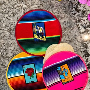 K&ouml;nnte beinhalten: Drei runde, farbenfrohe Stoffuntersetzer mit mexikanischem Serape-Design. Jeder Untersetzer zeigt ein anderes Loteria-Kartenbild: El Borracho, La Rosa und La Borracha. Die Untersetzer sind auf einer grauen Arbeitsplatte mit einem Stapel Tortillas im Hintergrund angeordnet.