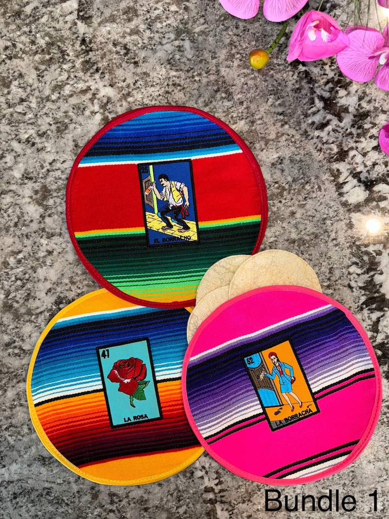 K&ouml;nnte beinhalten: Drei runde, farbenfrohe Stoffuntersetzer mit mexikanischem Serape-Design. Jeder Untersetzer zeigt ein anderes Loteria-Kartenbild: El Borracho, La Rosa und La Borracha. Die Untersetzer sind auf einer grauen Arbeitsplatte mit einem Stapel Tortillas im Hintergrund angeordnet.