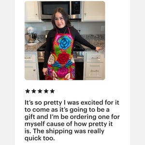 May include: A woman wearing a colorful floral apron with a black turtleneck shirt. The apron has a variety of flowers in shades of pink, red, blue, green, and yellow. The apron is made of a soft, woven fabric.