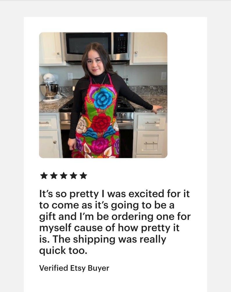 May include: A woman wearing a colorful floral apron with a black turtleneck shirt. The apron has a variety of flowers in shades of pink, red, blue, green, and yellow. The apron is made of a soft, woven fabric.