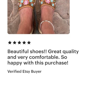 May include: A pair of brown leather sandals with floral embroidery in pink, blue, and yellow. The sandals have a light-colored sole and are worn on a stone surface. The text "Beautiful shoes!! Great quality and very comfortable. So happy with this purchase!" is also visible.