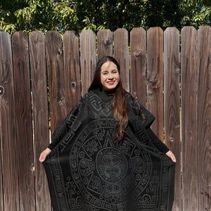 May include: A black poncho with a detailed Aztec calendar design. The poncho has fringe along the bottom and is worn by a person with long brown hair.