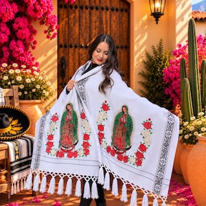 May include: A white rectangular shawl featuring two embroidered images of the Virgin of Guadalupe surrounded by red and white roses. The shawl has black geometric patterned borders and long white fringe along the bottom edge of the fabric.