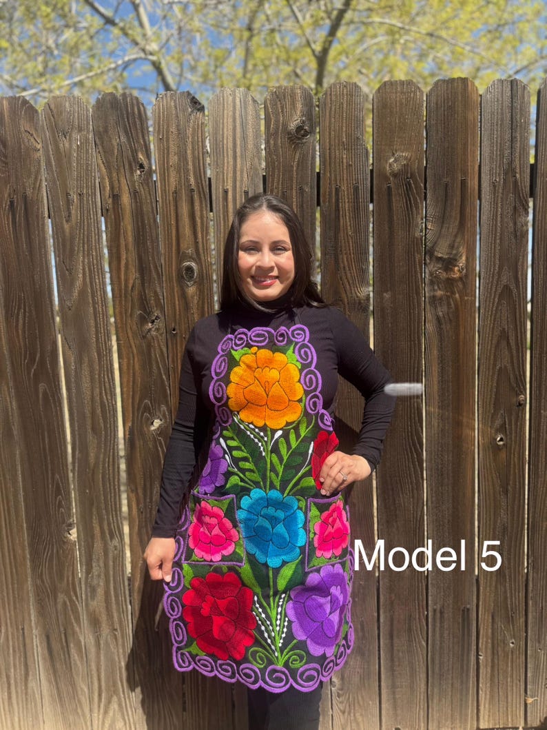 May include: A black apron with colorful embroidered flowers. The apron has a purple trim and two pockets. The model is wearing a black turtleneck shirt. The text "Model 5" is visible in the image.