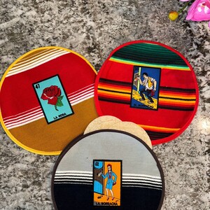 K&ouml;nnte beinhalten: Set aus drei runden, bunten Stoffuntersetzern mit mexikanischem Design. Jeder Untersetzer zeigt eine andere Loteria-Karte mit einer bunten Illustration und Text. Die Karten sind "La Rosa", "El Borracho" und "La Borracha".