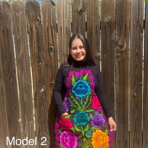 May include: A black apron with colorful embroidered flowers. The apron has a large pocket in the front and a pink trim around the edges. The model is wearing a black mock neck shirt and jeans. "Model 2" is written in the bottom left corner of the image.
