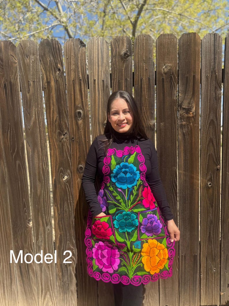May include: A black apron with colorful embroidered flowers. The apron has a large pocket in the front and a pink trim around the edges. The model is wearing a black mock neck shirt and jeans. "Model 2" is written in the bottom left corner of the image.