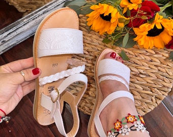 May include: White leather sandals with floral embossed detailing on the straps. The sandals have a braided strap and a buckle closure. The image also shows a beaded ankle bracelet and sunflowers.