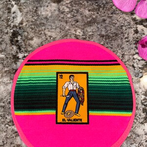 K&ouml;nnte beinhalten: Ein runder, rosa Topflappen mit einem farbenfrohen Serape-Muster. In der Mitte befindet sich eine gelbe Karte mit einer Comic-Abbildung eines Mannes, der ein Messer und einen Serape h&auml;lt. Die Karte tr&auml;gt die Aufschrift "12 El Valiente".