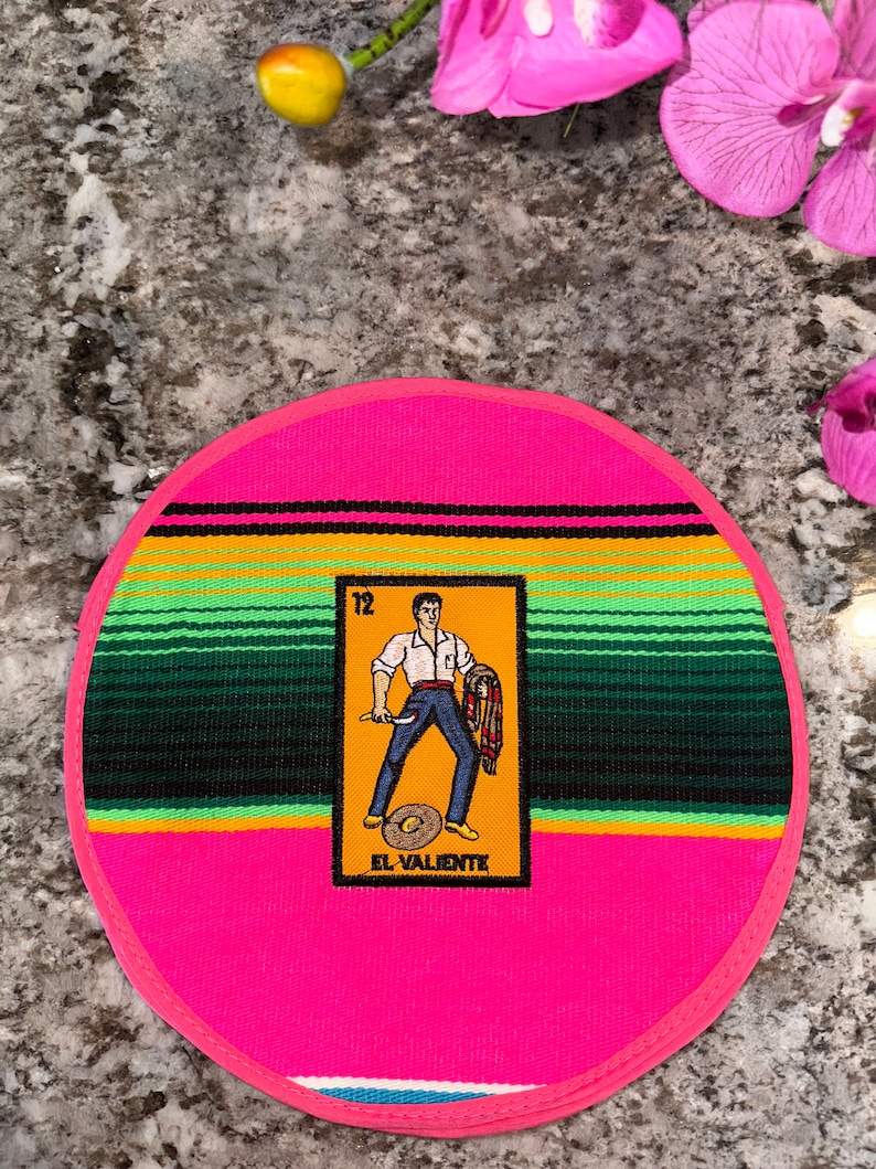 K&ouml;nnte beinhalten: Ein runder, rosa Topflappen mit einem farbenfrohen Serape-Muster. In der Mitte befindet sich eine gelbe Karte mit einer Comic-Abbildung eines Mannes, der ein Messer und einen Serape h&auml;lt. Die Karte tr&auml;gt die Aufschrift "12 El Valiente".