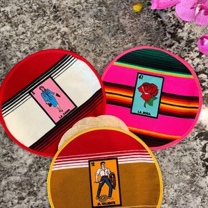 K&ouml;nnte beinhalten: Drei runde, farbenfrohe Stoffuntersetzer mit einem roten, wei&szlig;en und schwarzen Rand. Jeder Untersetzer zeigt ein anderes Loteria-Kartenbild. Der erste Untersetzer hat eine rosa Karte mit einer Frau in einem blauen Kleid und dem Text "La Dama". Der zweite Untersetzer hat eine rosa Karte mit einer roten Rose und dem Text "La Rosa". Der dritte Untersetzer hat eine gelbe Karte mit einem Mann, der ein Schwert h&auml;lt, und dem Text "El Valiente". Der Text "Bundle 3" ist in der rechten unteren Ecke des Bildes sichtbar.