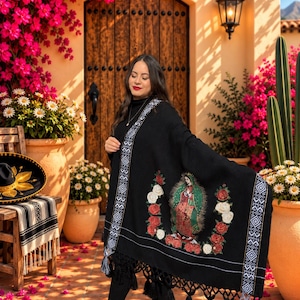 May include: A black shawl featuring an embroidered image of the Virgin of Guadalupe surrounded by red and white roses. The shawl has a white geometric patterned border along the edges and is finished with long black fringe tassels at the bottom hem.