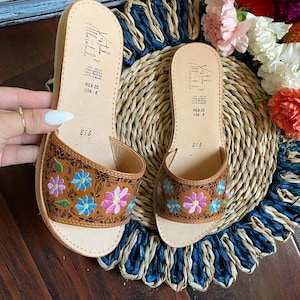 May include: A pair of brown leather sandals with floral embroidery in pink, blue, and purple. The sandals have a natural leather footbed and a small wedge heel. The text "Vestida" and "MEX-25 USA-8" is visible on the sandals.