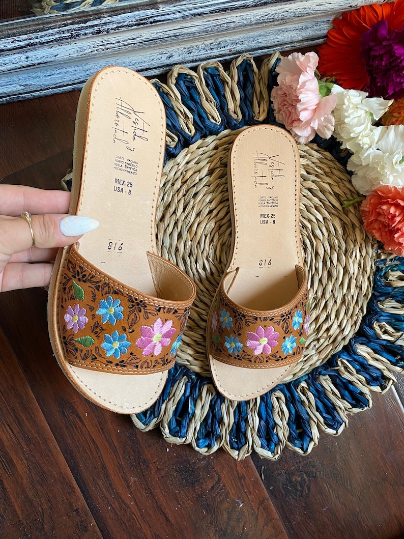 May include: A pair of brown leather sandals with floral embroidery in pink, blue, and purple. The sandals have a natural leather footbed and a small wedge heel. The text "Vestida" and "MEX-25 USA-8" is visible on the sandals.