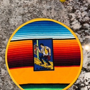 K&ouml;nnte beinhalten: Ein runder Untersetzer mit gelbem Rand und einem farbenfrohen Serape-Muster. In der Mitte befindet sich eine Karte mit einem Mann, der eine Flasche h&auml;lt, und dem Text "EL BORRACHO".