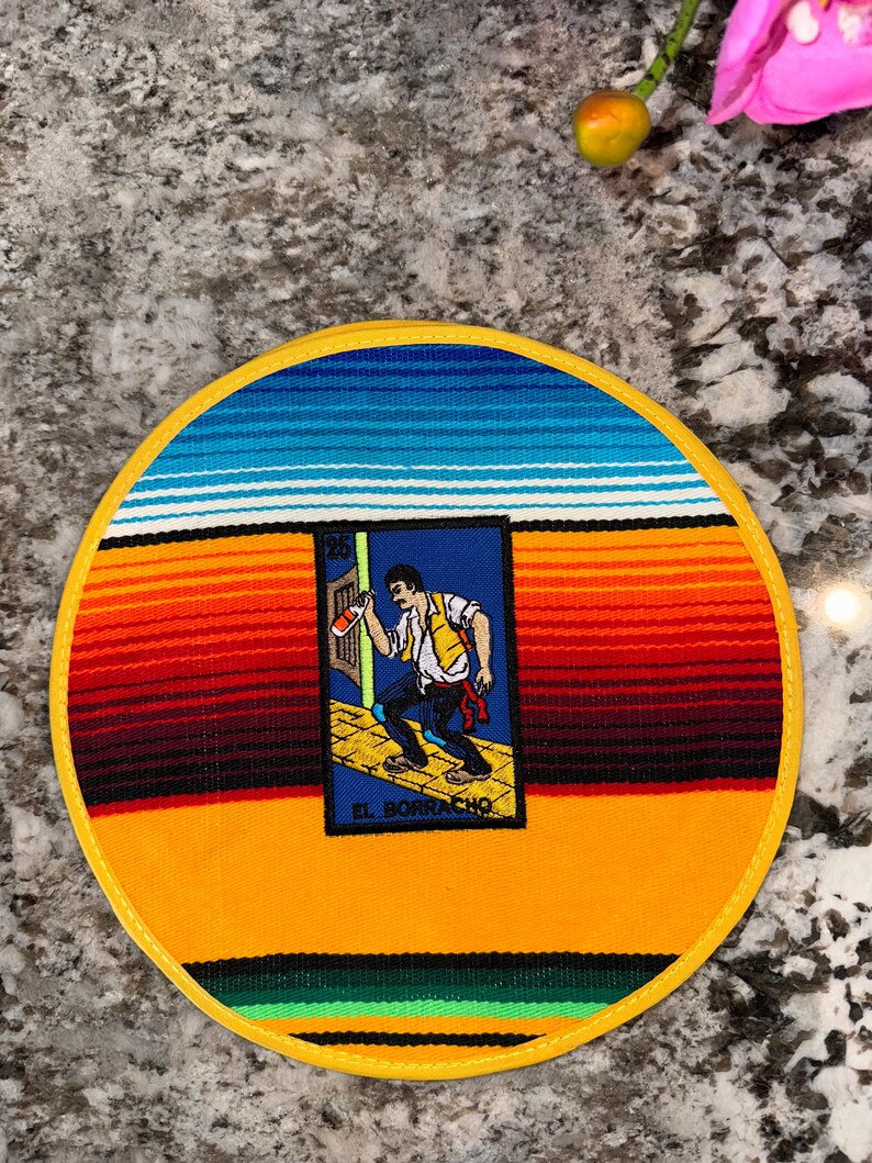 K&ouml;nnte beinhalten: Ein runder Untersetzer mit gelbem Rand und einem farbenfrohen Serape-Muster. In der Mitte befindet sich eine Karte mit einem Mann, der eine Flasche h&auml;lt, und dem Text "EL BORRACHO".