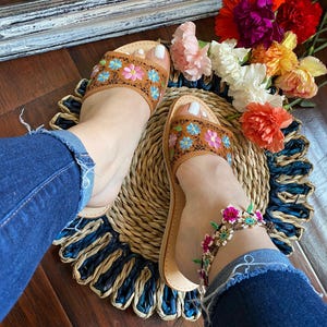 May include: Brown leather sandals with floral embroidery in pink, blue, and yellow. The sandals are worn with a beaded ankle bracelet featuring pink flowers. The sandals are on a woven mat with a blue and natural color scheme.