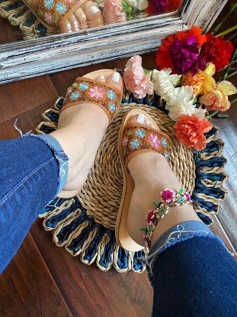 May include: Brown leather sandals with floral embroidery in pink, blue, and yellow. The sandals are worn with a beaded ankle bracelet featuring pink flowers. The sandals are on a woven mat with a blue and natural color scheme.