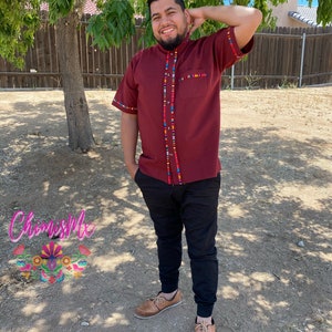 May include: A maroon short-sleeved shirt with colorful trim around the collar, cuffs, and down the front. The shirt is worn by a person with a beard and a smile. The person is standing in front of a wooden fence and a tree.