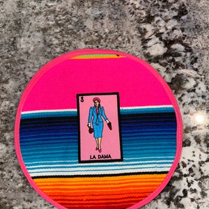 K&ouml;nnte beinhalten: Ein runder rosa Untersetzer mit einem bunten Streifenmuster und einer Karte mit einer Frau in einem blauen Kleid und dem Text "LA DAMA".