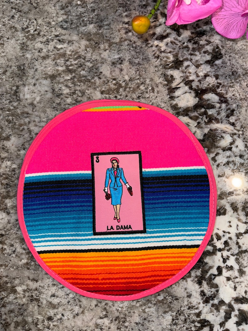 K&ouml;nnte beinhalten: Ein runder rosa Untersetzer mit einem bunten Streifenmuster und einer Karte mit einer Frau in einem blauen Kleid und dem Text "LA DAMA".