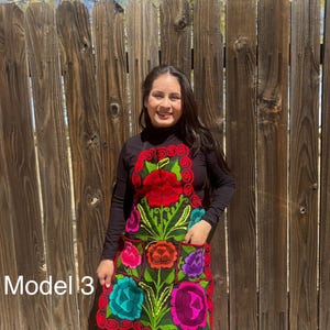 May include: A black apron with colorful embroidered flowers. The apron has pockets and a ruffled bottom. The text "Model 3" is visible in the image.