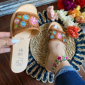 May include: Brown leather sandals with floral embroidery in blue, pink, and green. The sandals are open-toed and feature a woven sole. The image also shows a floral anklet and a colorful flower arrangement.