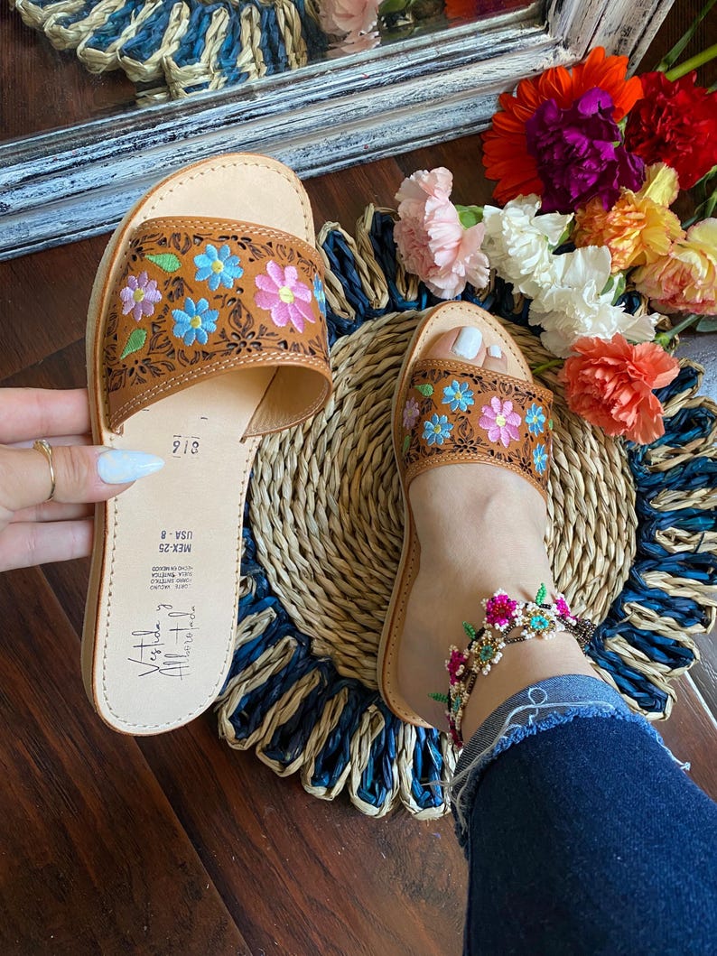 May include: Brown leather sandals with floral embroidery in blue, pink, and green. The sandals are open-toed and feature a woven sole. The image also shows a floral anklet and a colorful flower arrangement.