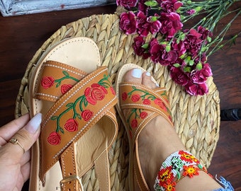 May include: A pair of brown leather sandals with a floral embroidery design. The sandals have a criss-cross strap design and an ankle strap with a buckle closure. The embroidery features red roses and green leaves.