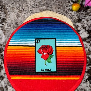 K&ouml;nnte beinhalten: Ein runder, rot-wei&szlig;-blauer gewebter Untersetzer mit rotem Rand. In der Mitte des Untersetzers befindet sich eine Loteria-Karte mit einer roten Rose und dem Text "La Rosa" und der Nummer "41".