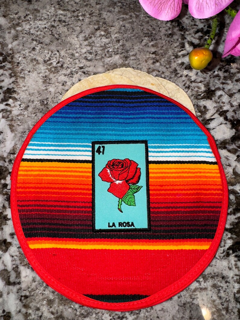 K&ouml;nnte beinhalten: Ein runder, rot-wei&szlig;-blauer gewebter Untersetzer mit rotem Rand. In der Mitte des Untersetzers befindet sich eine Loteria-Karte mit einer roten Rose und dem Text "La Rosa" und der Nummer "41".