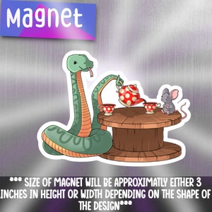 May include: A green snake pours tea from a red teapot with white polka dots into a teacup for a grey mouse. The snake and mouse are sitting at a wooden table with a spool base. The magnet is approximately 3 inches in height or width.
