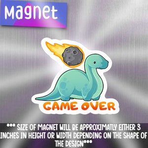 May include: A green dinosaur with a long neck is being hit by a gray asteroid with a yellow flame trail. The text "GAME OVER" is below the dinosaur. The magnet is approximately 3 inches in height or width.