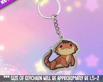 May include: A clear acrylic keychain featuring a cartoon bearded dragon with a small brown insect on its head. The keychain is attached to a silver metal ring and chain. The keychain measures approximately 1.5 to 2 inches in height or width.